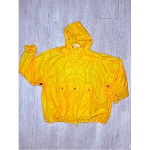 May include: A bright yellow pullover windbreaker jacket featuring a hood, a half-zip front closure, long sleeves with adjustable snap button cuffs, and a large rectangular front pocket with three visible snap buttons and a drawstring hem.