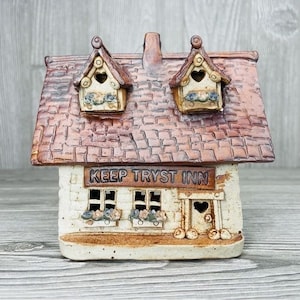 Może przedstawiać: Ceramiczny budynek w stylu wiejskiego domku z brązowym dachem i białymi ścianami. Budynek ma dwa okna z doniczkami na kwiaty i znak z napisem "Keep Tryst Inn".