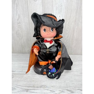 May include: A doll dressed as a vampire wearing a black cape with an orange lining, a black jacket, a white shirt, and a red bow tie. The doll has curly black hair and holds a small black bat-shaped basket with purple and green accents.