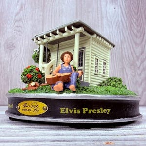 May include: A decorative figurine depicting a small house with a porch, a figure playing a guitar, and the text "Elvis Presley." The base includes a gold-coloured oval with the text "Elvis Park, Tupelo, MS."