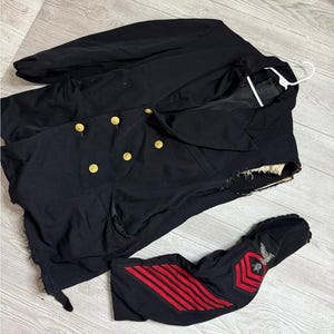 May include: A dark navy blue military jacket with gold buttons and a matching sleeve with red stripes and an emblem. The jacket is lying on a light gray wooden surface.