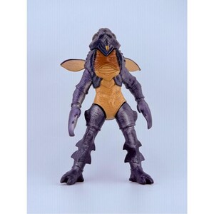 May include: A standing insectoid action figure with a dark purple exoskeleton, a tan chest plate, and a yellow pointed mouthpart. The figure features two small tan wings on its back, claw-like hands, and textured legs with fin-like protrusions.