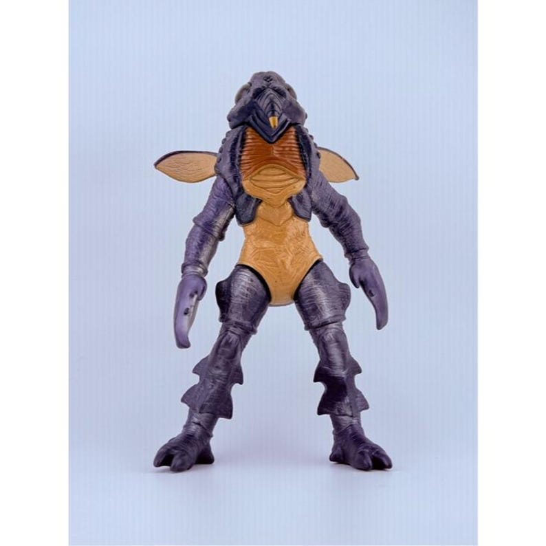 May include: A standing insectoid action figure with a dark purple exoskeleton, a tan chest plate, and a yellow pointed mouthpart. The figure features two small tan wings on its back, claw-like hands, and textured legs with fin-like protrusions.