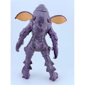 May include: A purple creature action figure with a textured, scaly body, two large tan ears with visible veining, and claw-like hands. The figure stands in an upright position, showcasing a detailed, rugged surface texture across its entire form.