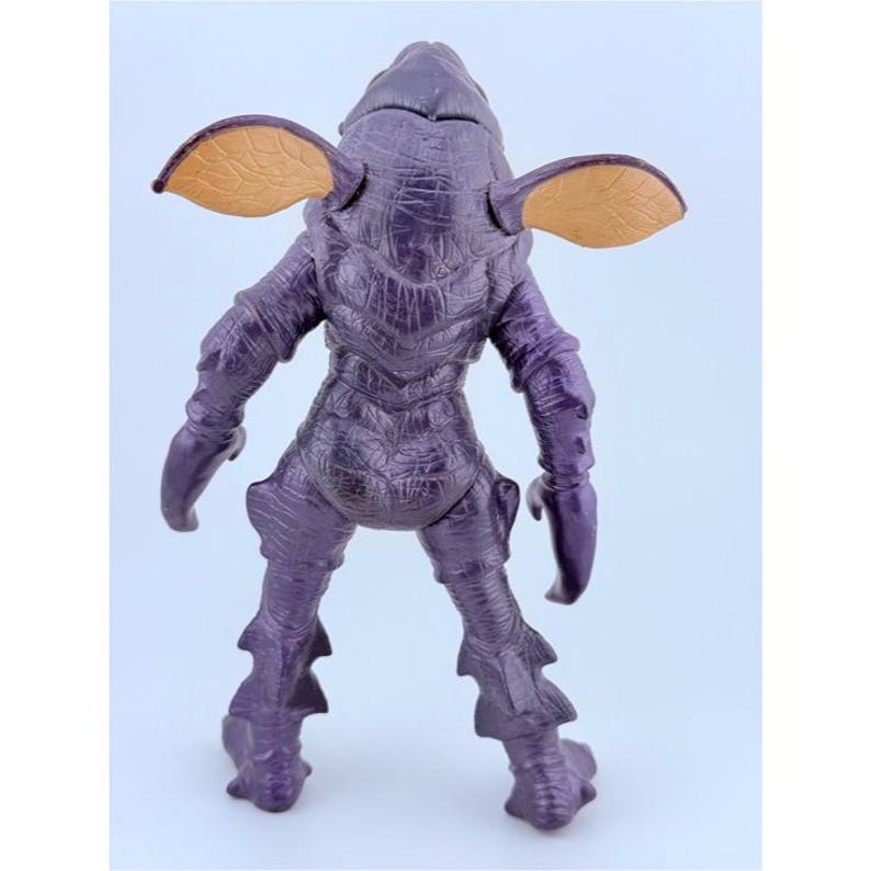 May include: A purple creature action figure with a textured, scaly body, two large tan ears with visible veining, and claw-like hands. The figure stands in an upright position, showcasing a detailed, rugged surface texture across its entire form.