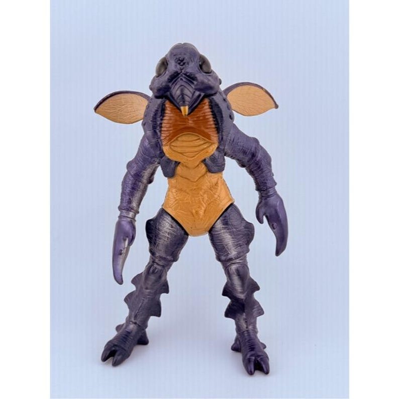 May include: A purple and tan insectoid action figure with a textured exoskeleton, large dark eyes, two small wings extending from the shoulders, and claw-like hands. The creature features a segmented torso, pointed feet, and a rigid, upright posture.