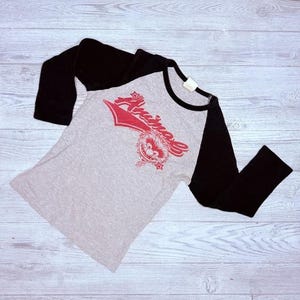 May include: A gray raglan-style t-shirt with black sleeves. The shirt features the word "Animal" in red script with a cartoon character graphic. The shirt is made of a soft material and is laid flat on a light wood surface.