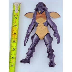 May include: A purple and tan insectoid action figure with large dark eyes, small wings, and claw-like hands. A yellow Stanley measuring tape is positioned vertically next to the figure, showing a height of approximately 7.5 inches for the toy.