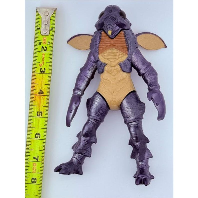 May include: A purple and tan insectoid action figure with large dark eyes, small wings, and claw-like hands. A yellow Stanley measuring tape is positioned vertically next to the figure, showing a height of approximately 7.5 inches for the toy.