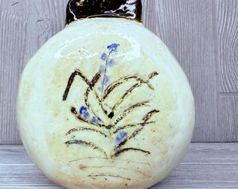 May include: A handmade ceramic vase with a rounded body and a dark brown rim. The vase is off-white with a floral design in brown and blue. The vase is a decorative item.