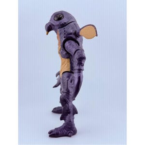 May include: A purple creature action figure with a textured, wrinkled skin surface, a large black eye, a pointed beak, and a tan-colored chest panel. The figure features articulated arms with claw-like hands, small fin-like protrusions on the legs, and a tan ear-like appendage extending from the side of the head.