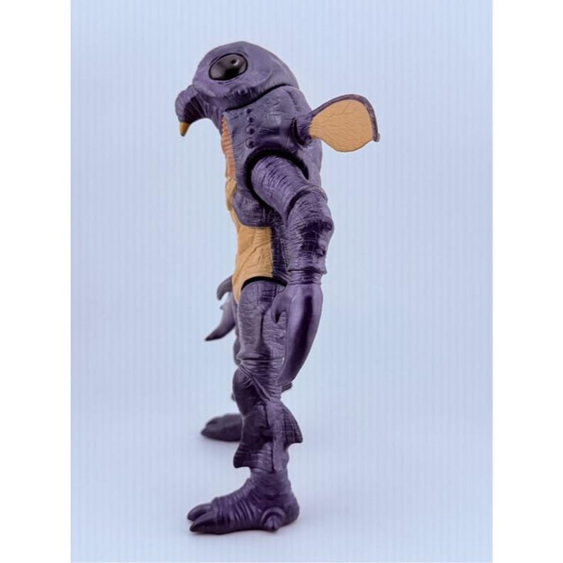May include: A purple creature action figure with a textured, wrinkled skin surface, a large black eye, a pointed beak, and a tan-colored chest panel. The figure features articulated arms with claw-like hands, small fin-like protrusions on the legs, and a tan ear-like appendage extending from the side of the head.