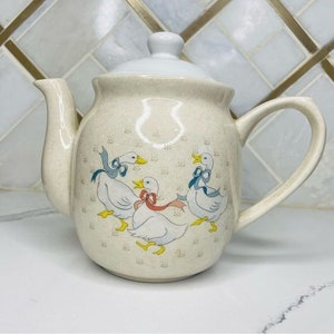 May include: A cream-colored ceramic teapot with a white lid and a handle. The teapot is decorated with a pattern of three white geese with blue and pink bows. The geese are surrounded by a light brown and white floral pattern.