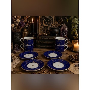 Op de afbeelding: Een set van vier diepblauwe keramische theekopjes met bijpassende schoteltjes, elk versierd met gouden hemelse ster- en maanpatronen. De kopjes hebben gebogen oren en de schoteltjes hebben een wit midden met een gouden steraccent en gouden randen.