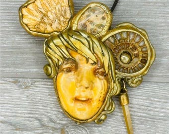 Pode incluir: Um colar com um pingente dourado que apresenta um rosto esculpido detalhado com uma coroa de desenhos intrincados. O pingente está preso a um cordão preto.