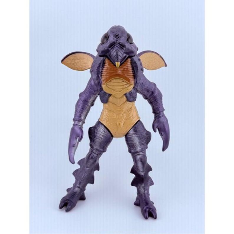 May include: A purple and tan insectoid creature action figure with a textured exoskeleton, large dark eyes, a pointed snout, and two small tan wings extending from its back. The figure features claw-like hands and segmented legs with pointed feet.