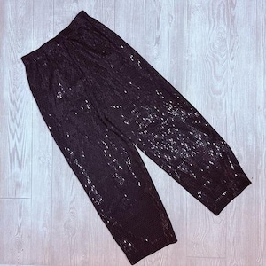 Puede incluir: Pantalones negros con lentejuelas y cintura elástica. Los pantalones están hechos de una tela oscura con lentejuelas plateadas dispersas, creando un efecto brillante. Los pantalones están sobre una superficie clara con vetas de madera.