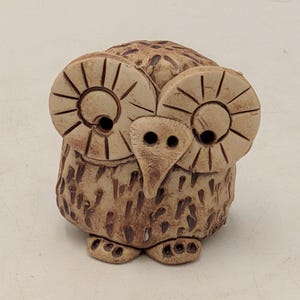 May include: A small, hand-sculpted ceramic owl figurine in a light beige tone. The owl features two large, circular eyes with etched radial lines, a pointed beak, and a textured body decorated with dark brown vertical dashes and small feet.