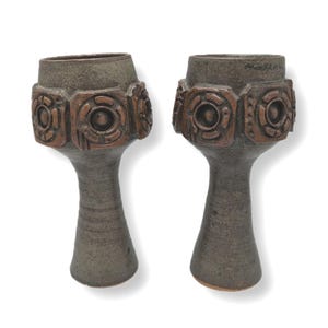 May include: Two ceramic vases with a dark grey speckled finish. Each vase has a flared top and a decorative band of geometric shapes in a copper colour. The vases have a slender, elongated stem.