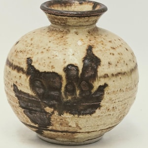 May include: A small, round ceramic vase with a speckled beige finish and dark brown abstract design. The vase has a narrow neck and a slightly flared rim. The design appears to be a stylized landscape or silhouette.