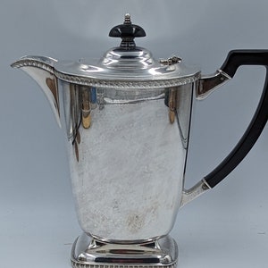 May include: A silver teapot with a black handle and a black knob on the lid. The teapot has a decorative design around the base and lid.