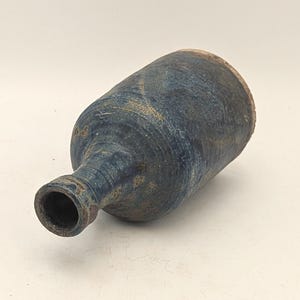 May include: A handmade ceramic bottle with a dark blue glaze and a textured surface. The bottle has a cylindrical body and a narrow neck. The rim of the bottle is a lighter brown colour. The bottle is approximately 20 cm long.