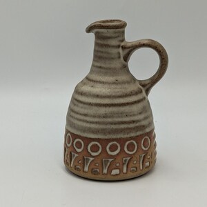 May include: A small, brown and white ceramic jug with a handle. The jug has a brown and white striped design and a brown base with a white geometric pattern.