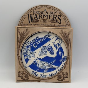 May include: A ceramic bread warmer featuring a blue and white design of the state of North Carolina with a cardinal bird. The text "North Carolina" and "The Tar Heel State" are printed on the warmer. The warmer is packaged in a brown cardboard box with wheat stalks and the text "Bread & Bun Warmers".