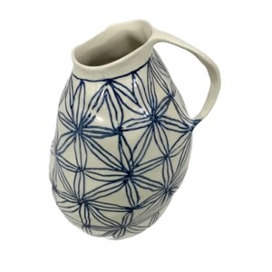 May include: A ceramic jug with a white base and a blue floral pattern. The jug has a curved handle and a wide opening. The design is a repeating pattern of stylized flowers. The jug is likely handmade and suitable for serving beverages.