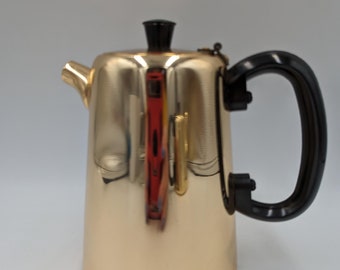 Op de afbeelding: Een gouden metalen koffiekan met een zwarte handgreep en deksel. De kan heeft een glanzend, reflecterend oppervlak.