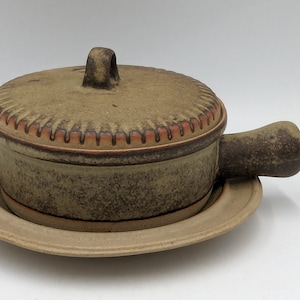 May include: A brown ceramic casserole dish with a lid and a handle. The dish has a textured surface and a brown and orange rim.