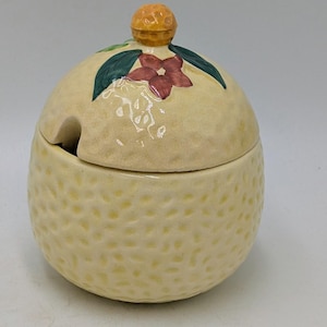 May include: A light yellow ceramic sugar bowl with a lid. The lid is decorated with a red flower, green leaves, and an orange knob. The bowl has a textured surface and a small opening for a spoon.