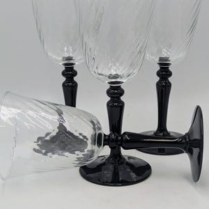 May include: Four clear wine glasses with a textured design on the bowl, set on black stems and bases. One glass is lying on its side. The glasses are arranged against a white background.