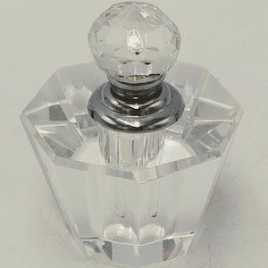 May include: A clear glass perfume bottle with a faceted, spherical stopper. The bottle has a geometric, multi-sided shape and a silver-coloured metal collar. The stopper is clear and reflects light, adding elegance to the design.
