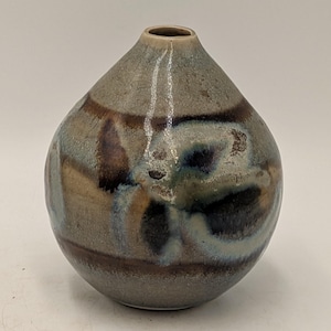 May include: A small, round ceramic vase with a narrow neck and a tapered opening. The surface features a mottled glaze in shades of muted grey, earthy brown, and deep blue, with swirling patterns that create a textured, multi-tonal appearance.