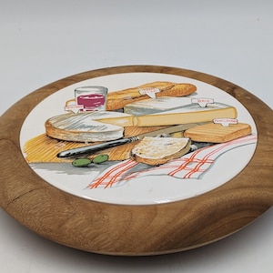 May include: A round wooden cheese board with a white ceramic insert featuring a painted illustration of a cheese plate with various cheeses, bread, olives, and a glass of wine. The ceramic insert has the words "Brie", "Pont L'Eveque", and "Cantal", printed on it.
