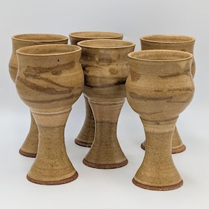 May include: Six handmade ceramic goblets with a speckled tan and brown glaze. The goblets have a bulbous bowl, a slender stem, and a flared base. The pottery has a rustic, earthy aesthetic.