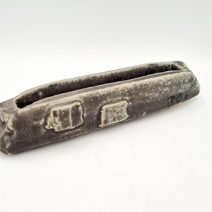 May include: A long, narrow, grey ceramic planter with a rough, textured surface. The planter has a rectangular opening in the centre and a small, rectangular indentation on the side. The planter is marked with the number "301".