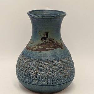 May include: A handmade ceramic vase. The lower half is textured and dark blue, while the upper section is smooth and lighter blue. It has a distinctive, organic form and a dark brown band near the top. A decorative piece for the home.