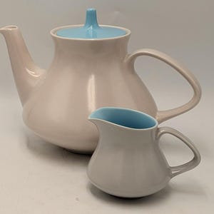 May include: A light pink teapot with a smooth finish and a light blue lid featuring a central knob, accompanied by a matching light pink creamer pitcher that has a light blue interior. Both pieces have curved handles and a rounded, bulbous shape.