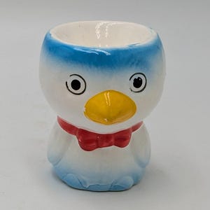 May include: A white ceramic egg cup shaped like a penguin with a blue and white body, yellow beak, and red bow tie.