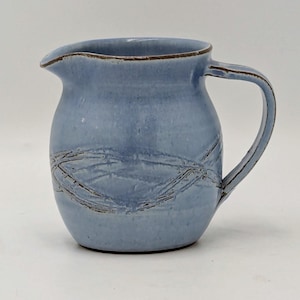 May include: A light blue ceramic jug with a curved handle and spout. The jug has a textured, crisscross pattern around the body and a brown rim. This handmade jug is suitable for serving drinks. The height is approximately 15 cm.