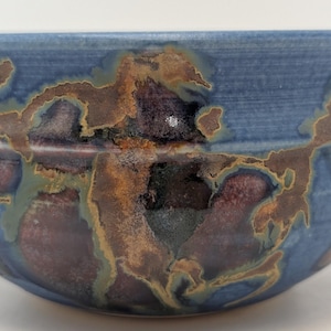 May include: A handcrafted ceramic bowl, featuring a blue exterior and abstract brown, green, and purple patterns. The bowl has an irregular shape and a textured surface, enhancing its unique, artistic appeal.