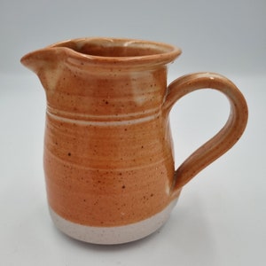 May include: A small, hand-thrown ceramic jug with a speckled orange glaze and a white rim. The jug has a rounded body and a small handle.
