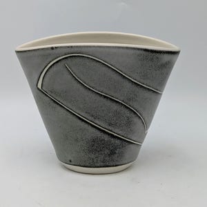 May include: A grey ceramic vase with a distinctive shape and a white interior. The vase has a textured surface and white curved lines. The base and rim are white. This decorative vase is suitable for home decor.