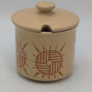 May include: A beige ceramic sugar bowl with a matching lid and a small knob. The bowl features a repeating sunburst design in a darker beige colour. The lid has a small notch for a spoon.