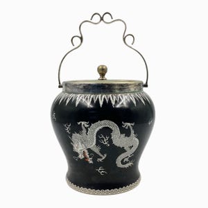 May include: A black ceramic container with a silver lid and handle. The container features a white dragon design and decorative silver trim. The handle has a unique, ornate shape. A small, gold-coloured knob sits atop the lid.