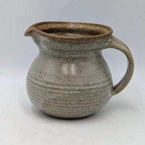 May include: A handmade stoneware creamer with a rounded body and a curved handle. The creamer has a spout for easy pouring and a light brown rim. The exterior is a neutral grey with subtle horizontal lines.