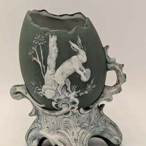 May include: A sage green egg-shaped vessel with a jagged top edge, featuring white relief carvings of a rabbit and a tree. The vessel is supported by an ornate, light-coloured base with scrollwork details and includes a small handle on the side.