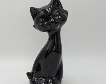 May include: A glossy black ceramic cat ornament. The cat is sitting upright with its head turned slightly to the side. The ornament has a sleek, minimalist design, with smooth curves and a polished finish. The cat's features are subtly defined.